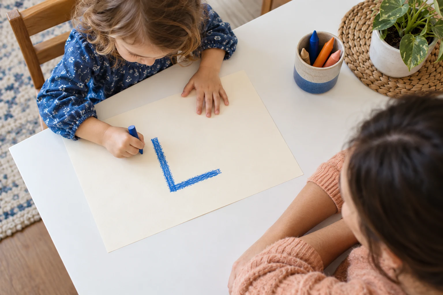 From above, a child at the upper left uses a broad blue crayon beside a large L while an adult observes from the lower right.